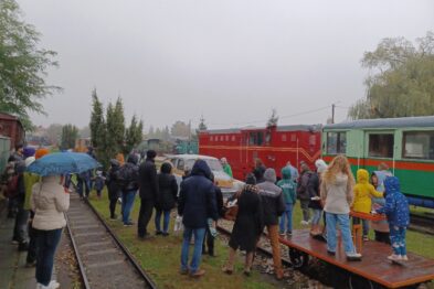 Grupa osób stoi na peronie przy czerwonym parowozie i zielonych wagonach; niektórzy trzymają parasole. Towarzyszą im platforma kolejowa oraz tory. Pogoda wydaje się być pochmurna i deszczowa.