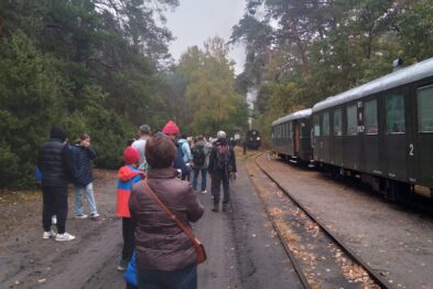 Grupa ludzi zgromadziła się na peronie obok pociągu retro, który stoi na torach w otoczeniu drzew. Osoby są ubrane w ciepłe kurtki, niektóre z nich mają na głowach czapki, sugerując chłodną porę roku. Wagony pociągu są zielone i wydaje się, że są wykonane z metalu.
