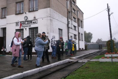 Grupa osób stoi na peronie przed budynkiem z napisem 