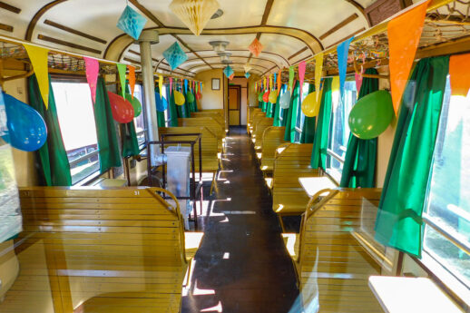 Wagon kolejowy jest udekorowany kolorowymi balonami i girlandami, tworząc przytulną atmosferę na uroczystość. Drewniane ławki ustawione są wzdłuż pojazdu, a okna z zielonymi zasłonami wpuszczają światło do środka. Stolik na końcu wagonu jest przygotowany na przyjęcie z nakryciami i dekoracjami.