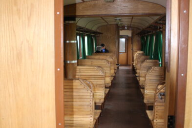 Wnętrze drewnianego wagonu kolejowego z zielonymi, tapicerowanymi siedzeniami ustawionymi parami wzdłuż obu długich ścian. Podłoga pokryta jest brązową wykładziną, a sufit zaopatrzony w okrągłe lampy. Drzwi na pierwszym planie są otwarte, pokazując korytarz prowadzący przez środek wagonu.