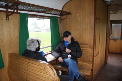 Dwóch mężczyzn w historycznym wagonie kolejowym - jeden siedzi przy drewnianym oknie z mikrofonem, drugi operuje kamerą. Wnętrze wagonu jest utrzymane w starym stylu z drewnianymi ławkami i zielonymi zasłonkami. Za oknem widać zielony krajobraz i częściowo widoczne budynki zewnętrzne.