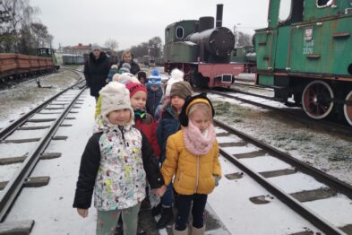 Grupa dzieci stoi na zewnątrz między torami kolejowymi, dwie stare, zielone lokomotywy parowe znajdują się w tle. Na ich twarzach widać różne emocje, dzieci są ubrane w ciepłe zimowe ubrania. Niebo jest pochmurne, a podłoże pokrywa cienka warstwa śniegu.