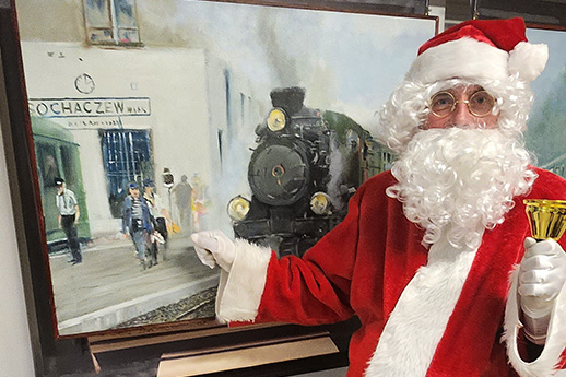 Osoba przebrana za Świętego Mikołaja w czerwonym kostiumie z białą brodą i czapką pozująca obok obrazu przedstawiającego stację kolejową i parowóz. Mikołaj trzyma w ręku złoty dzwoneczek. Tło fotografii to część wnętrza, w którym dominuje barwa biała oraz elementy dekoracyjne.