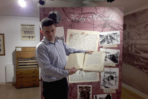The man in a light shirt and dark trousers is holding an open, old book in his hands. He is in a room with a museum exhibition, where an educational board on railway topics is placed on the wall. In the background, there are archival photos and documents related to the railway, as well as a wooden shelf with binders and cardboard boxes.