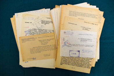 The set of documents is lying on a blue surface, containing carefully arranged, colorful sheets of paper, some of which bear stamps and handwritten annotations. Among the documents, there are technical descriptions and schematics, suggesting their connection to railway planning or operational activities. The documentation has a used appearance but is kept in good condition with historical value.