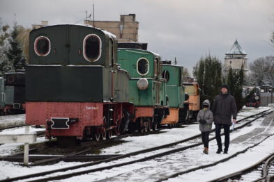 Starsza zielona lokomotywa parowa stoi na torach obok peronu pokrytego lekkim śniegiem; kilka innych wagonów i lokomotyw znajduje się w tle. Przed lokomotywą spacerują dwie osoby, dorosły i dziecko, trzymają się za ręce i mają na sobie ciepłe ubrania. W tle widoczne są także inne elementy infrastruktury kolejowej, w tym wieża ciśnień.