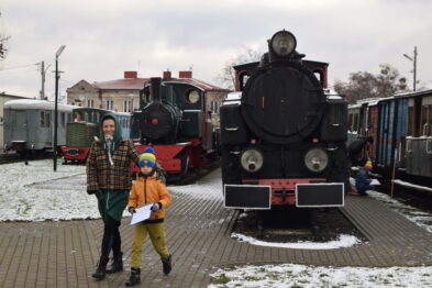 Starsza kobieta i młody chłopiec pozują do zdjęcia obok starego parowozu na stacji kolejowej. Chłopiec trzyma model pociągu i kartkę, a za nimi widać kolejne wagony i lokomotywy. Na peronie leży śnieg, a w tle widoczne są budynek stacyjny i drzewa.