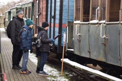 Troje osób stoi na peronie obok starego wagonu kolejowego, gdzie dorosły rozmawia z dzieckiem, a drugie dziecko spogląda w stronę drzwi wagonu. W tle widać kolejne zimowe pojazdy kolejowe i lekki śnieg na ziemi. Wszyscy są ubrani w ciepłe zimowe ubrania, co wskazuje na chłodną pogodę.
