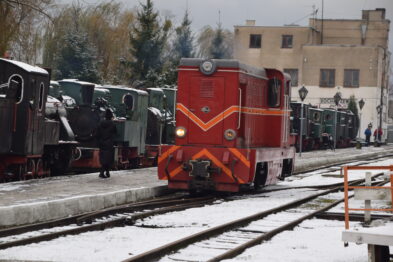 Czerwony spalinowy lokomotyw stoi na pierwszym planie torów pokrytych cienką warstwą śniegu. W tle widać inne lokomotywy i wagony ustawione na równoległych torach, tworzące ekspozycję. Nieduże płatki śniegu padają z szarego nieba, a otoczenie tworzy wrażenie chłodnej, zimowej aury.