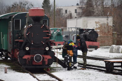 Stara lokomotywa parowa o czerwono-czarnym malowaniu stoi na torach obok zielonego wagonu; za nią widać inny, mniej wyraźny pojazd kolejowy. Dwóch mężczyzn i dziecko ubrane w ciepłe, zimowe ubrania stoją obok tej lokomotywy, wydają się być zaangażowani w rozmowę. Otoczenie jest wilgotne i pokryte cienką warstwą śniegu, a w tle widoczne są białe bloki kamienia oraz drzewa