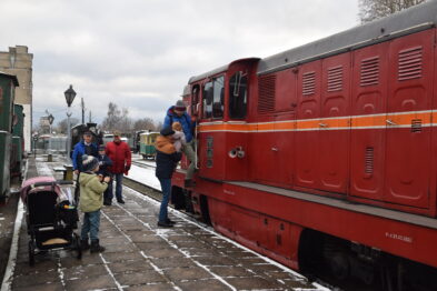 Czerwony lokomotywa stoi na torach obok peronu pokrytego śniegiem. Ludzie, w tym rodziny z dziećmi, oglądają pojazd i rozmawiają ze sobą, a niektóre dzieci wskazują na lokomotywę. W tle widać budynki stacyjne i latarnię uliczną.