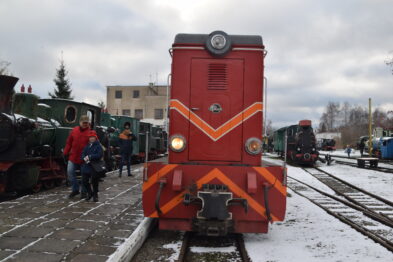 Czerwona lokomotywa stoi na torach kolejowych, przygotowana do jazdy z włączonymi światłami. W tle widać inne parowozy oraz grupę ludzi, którzy zgromadzili się na wydarzeniu. Śnieg pokrywa część ziemi oraz torowisko, co wskazuje na zimową porę roku.