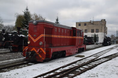 Czerwono-żółta lokomotywa stoi na torach obok peronu; w tle widać inne lokomotywy i wagony. Na ziemi leży cienka warstwa śniegu, a w powietrzu są widoczne płatki śniegu. Niebo jest pochmurne co sugeruje zimową aurę.