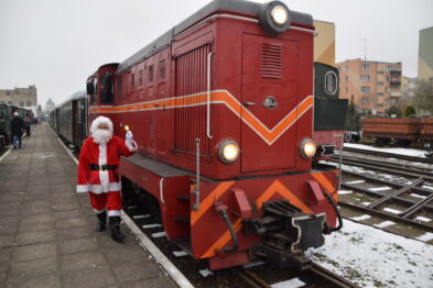 Czerwony wąskotorowy pociąg stoi na torach obok peronu, a obok niego pozuje postać przebrana za Świętego Mikołaja. Na ziemi widać niewielką ilość śniegu. W tle rozpoznajemy zabudowania miejskie w szarym, zimowym świetle.