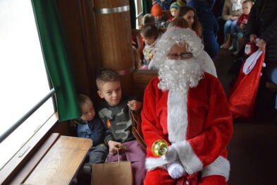 Osoba przebrana za Świętego Mikołaja siedzi w drewnianej wnęce wagonu kolejowego obok małych dzieci, trzymając dzwonek w prawej dłoni. Wagon jest wypełniony ludźmi, przeważnie dziećmi i ich opiekunami. W tle widoczne są inne wagony oraz okna, przez które przebija dzienny światło.