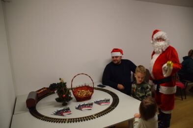 Osoba przebrana za Mikołaja stoi obok mężczyzny i dziecka siedzącego przy stole z makietą kolejki elektrycznej. Na stole widoczny jest także koszyk z przekąskami oraz mała choinka. Wszyscy na zdjęciu mają na głowach czerwone czapki świąteczne i wydają się być wesołego nastroju.