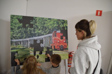 Kobieta i dwoje dzieci stoją przed dużym puzzlem z obrazem pociągu wąskotorowego przyklejonym do ściany. Dzieci współpracują nad układaniem brakujących elementów. W tle widoczny jest gaśnica i znak wyjścia ewakuacyjnego.