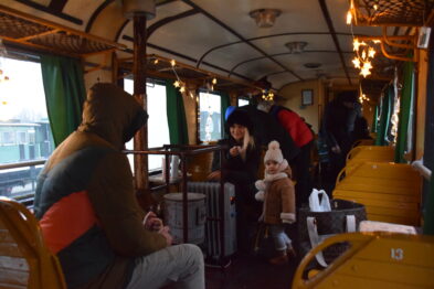 Wagon kolejki wąskotorowej jest wypełniony pasażerami; widać drewniane ławki i dekoracje świąteczne. Ludzie siedzą naprzeciwko siebie, w centrum kobieta trzyma dziecko na kolanach. Oświetlenie wnętrza daje ciepłe żółte światło, co tworzy przytulną atmosferę.