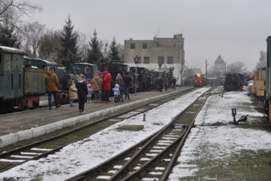 Grupa ludzi gromadzi się na peronie kolejowym przyozdobionym na świąteczne Mikołajki, przyglądając się lokomotywom i wagonom kolei wąskotorowej. Niebo jest zachmurzone, a na ziemi widać ślady śniegu, co nadaje scenerii zimowy charakter. Z tyłu zbliża się kolejna lokomotywa emitująca dym, co dodaje dynamizmu stacjonarnemu zestawowi pociągów.