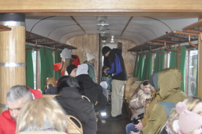 Ludzie zajmują miejsca w zabytkowym wagonie kolejowym, w którym zachowano oryginalny wystrój z drewnianymi ławkami i zielonymi zasłonkami. Osoba w czarnym nakryciu głowy chodzi między rzędami siedzeń, w tle widać okna i część wnętrza wagonu. Atmosfera jest świąteczna i rodzinna, pasażerowie w różnym wieku wydają się być zaangażowani w jakieś działanie lub rozmowy.