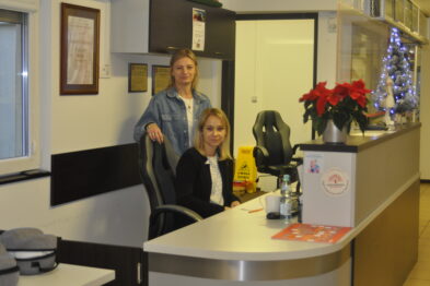 Dwie kobiety stoją za ladą informacyjną wewnątrz budynku. Na ladzie widać dekoracyjną czerwoną gwiazdę oraz małą choinkę świąteczną wyposażoną w oświetlenie. W tle znajduje się drzwi oraz plakat na ścianie.
