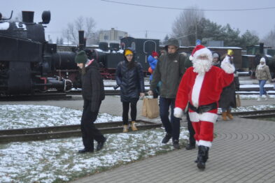 Postać w stroju Świętego Mikołaja idzie po torach kolejowych, a za nią można dostrzec grupę osób i stare lokomotywy. Na ziemi leży trochę śniegu, wskazując na zimową porę roku. W tle widoczna jest kolekcja zabytkowych parowozów i wagonów, co podkreśla tematykę kolejową miejsca.