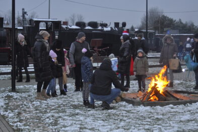 Grupa ludzi stoi na zewnątrz wokół ogniska, na ziemi widać lekki śnieg. W tle znajdują się lokomotywy i wagony kolejowe. Osoby są ubrane w ciepłe zimowe ubrania, a niebo jest zaciemnione, co wskazuje na późne popołudnie lub wczesny wieczór.