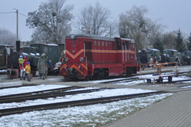 Czerwona lokomotywa staje na śnieżnej stacji obok peronu, przy której grupa osób w ciepłych ubraniach obserwuje otoczenie. Po obu stronach torów znajdują się kolejowe wagony i infrastruktura, a także śnieg delikatnie przykrywa ziemię. Widoczne są drzewa bez liści, a chmurne niebo wskazuje na zimową aurę.