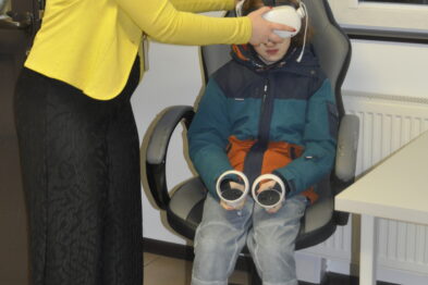 Kobieta ubrana w żółty sweter pomaga małemu chłopcu założyć białe gogle VR. Chłopiec siedzi na obrotowym krześle, ma na sobie kolorową kurtkę i brązowe buty. W tle widać białe drzwi i plakat z oknem.
