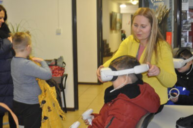 Dziecko siedzi na krześle z białym urządzeniem VR na głowie, a kobieta w żółtym płaszczu trzyma rękę na urządzeniu. W tle widoczna jest grupa osób, w tym dziecko z podobnym urządzeniem VR. Wszyscy znajdują się w pomieszczeniu z jasnym oświetleniem.
