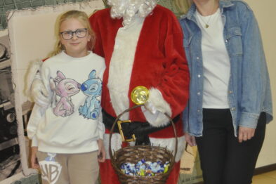 Osoba przebrana za Mikołaja stoi pomiędzy dwiema dziewczynkami uśmiechającymi się do kamery. Postać Mikołaja trzyma w ręku koszyk pełen słodyczy. W tle widoczne są dekoracje świąteczne i plakat z napisem „Świąteczne Warsztaty”.