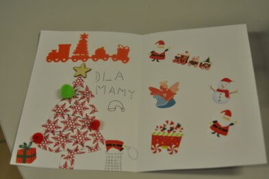 Otwarta kartka z życzeniami świątecznymi leży na płaskiej powierzchni, zdobiona ręcznie wykonanymi dekoracjami. Czerwona choinka z białymi śnieżynkami, pociąg i inne świąteczne symbole wypełniają stronę, z napisem 