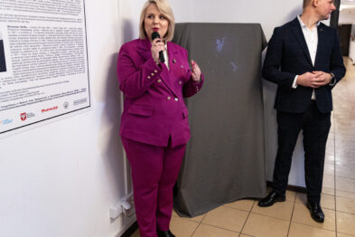 Kobieta w fioletowym garniturze trzyma mikrofon i coś wygłasza, stojąc obok osłoniętej szarej płachty. Obok niej stoi mężczyzna w ciemnym garniturze z rękami za plecami, który wydaje się być jej towarzyszącym. Na ścianie w tle widoczne są rozległe informacyjne teksty i grafiki.