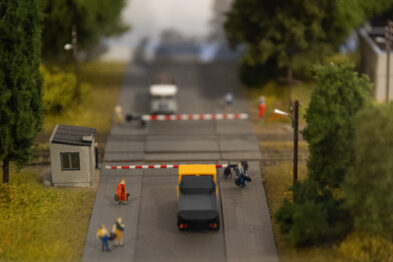 Zdjęcie przedstawia makietę kolejową z fragmentem miniaturowego przejazdu kolejowego z opuszczonymi szlabanami, przez które przejeżdża samochód. Wokół są miniaturowe postaci ludzkie oraz model domku, roślinności i drogowskazy. Całość wykonana jest z dużą dbałością o szczegóły imitujące rzeczywistość.