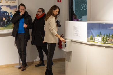 Trzy osoby oglądają ekspozycję w muzeum; dwie kobiety i mężczyzna. Jedna z kobiet uruchamia instalację za pomocą przycisku na białym postumencie. W tle widać model kolei zielono-białym krajobrazem i modele budynków.
