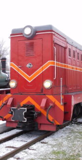 Czerwona lokomotywa wąskotorowa stoi na torach, a jej powierzchnia jest przykryta lekką warstwą śniegu. Obok lokomotywy widać inny zielony wagon kolejowy oraz ciąg kolejnych wagonów ciągnących się wzdłuż torów. Teren wokół torów pokrywa biały puch, a tło obrazu stanowi zimowe niebo.