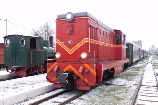 Czerwona lokomotywa wąskotorowa stoi na torach, a jej powierzchnia jest przykryta lekką warstwą śniegu. Obok lokomotywy widać inny zielony wagon kolejowy oraz ciąg kolejnych wagonów ciągnących się wzdłuż torów. Teren wokół torów pokrywa biały puch, a tło obrazu stanowi zimowe niebo.