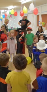 Grupa dzieci i dorosłych bawi się w jasnym pomieszczeniu udekorowanym kolorowymi balonami. Niektóre dzieci mają na głowach kolorowe czapki, a jedno z nich trzyma żółty model balonu. Animator w czarnym kapeluszu prowadzi zabawę, trzymając mikrofon i gestykulując do uczestników.