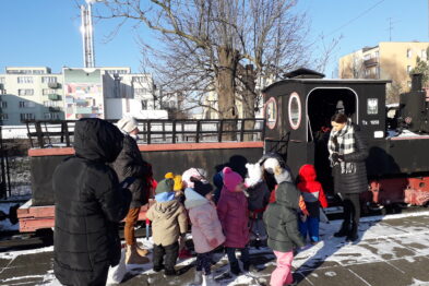 Wycieczka składająca się z dzieci i dwóch osób dorosłych zwiedzająca skansen. Widoczna w tle lokomotywa wąskotorowa.