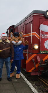 Przy czerwonej lokomotywie pozują mężczyzna w stroju sw. Mikołaja, wysoki mężczyzna w czerwonej świątecznej czapce i maskotka muzeum - maszynista Ryś