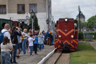 Muzealny pociąg z czerwona lokomotywą wjeżdża w peron, na którym stoją turyści